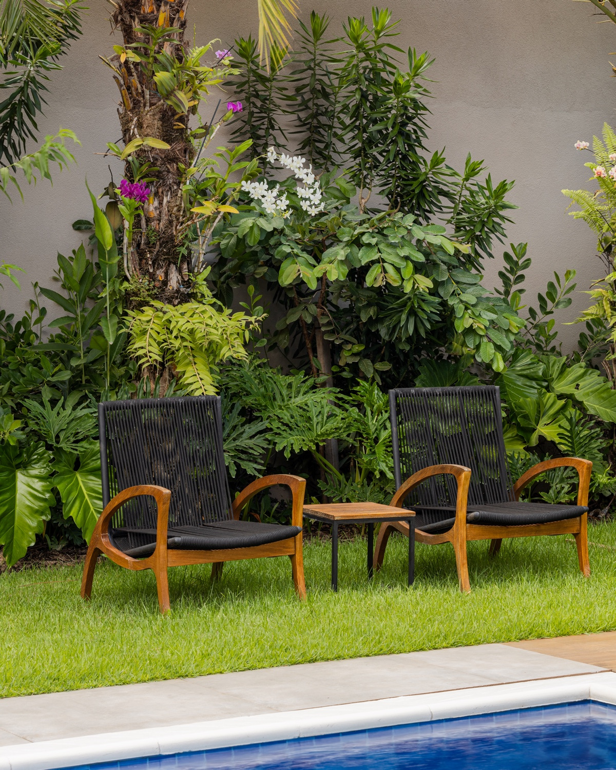 Duas poltronas de madeira e corda com mesinha lateral sobre o gramado à beira da piscina, diante de canteiro tropical com orquídeas roxas e brancas.
