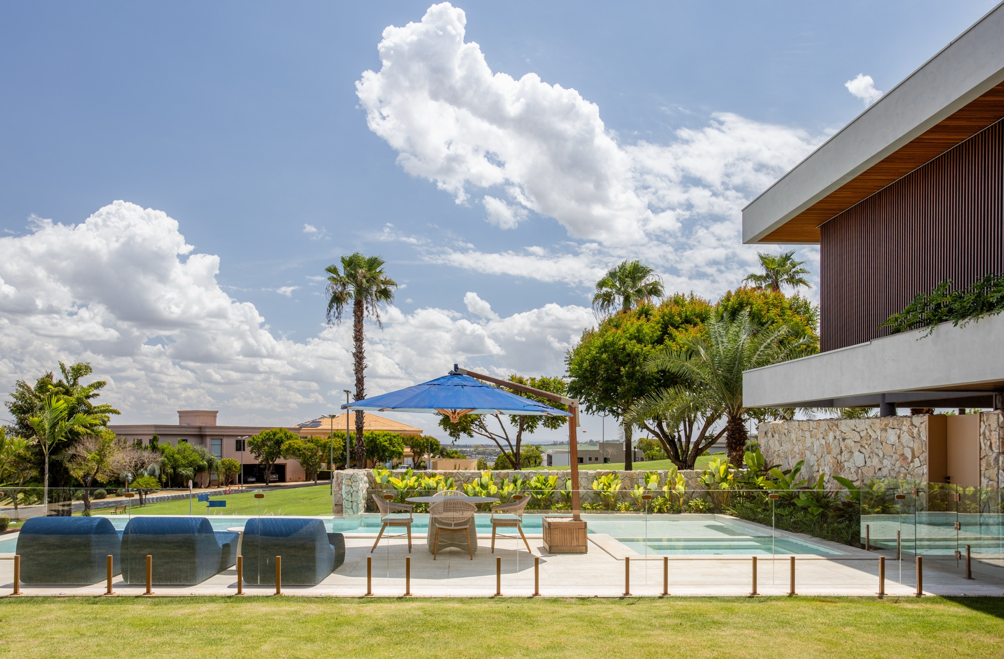 Piscina com guarda-sol azul e espreguiçadeiras, gramado verde em primeiro plano e palmeiras recortadas contra o céu de nuvens