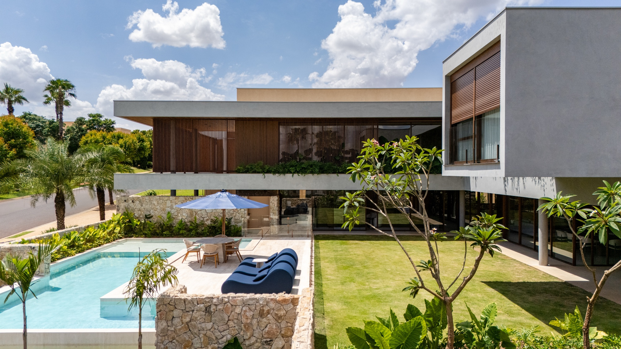 Piscina e gramado ao lado da casa, com árvore jovem em primeiro plano e guarda-sol azul junto à borda