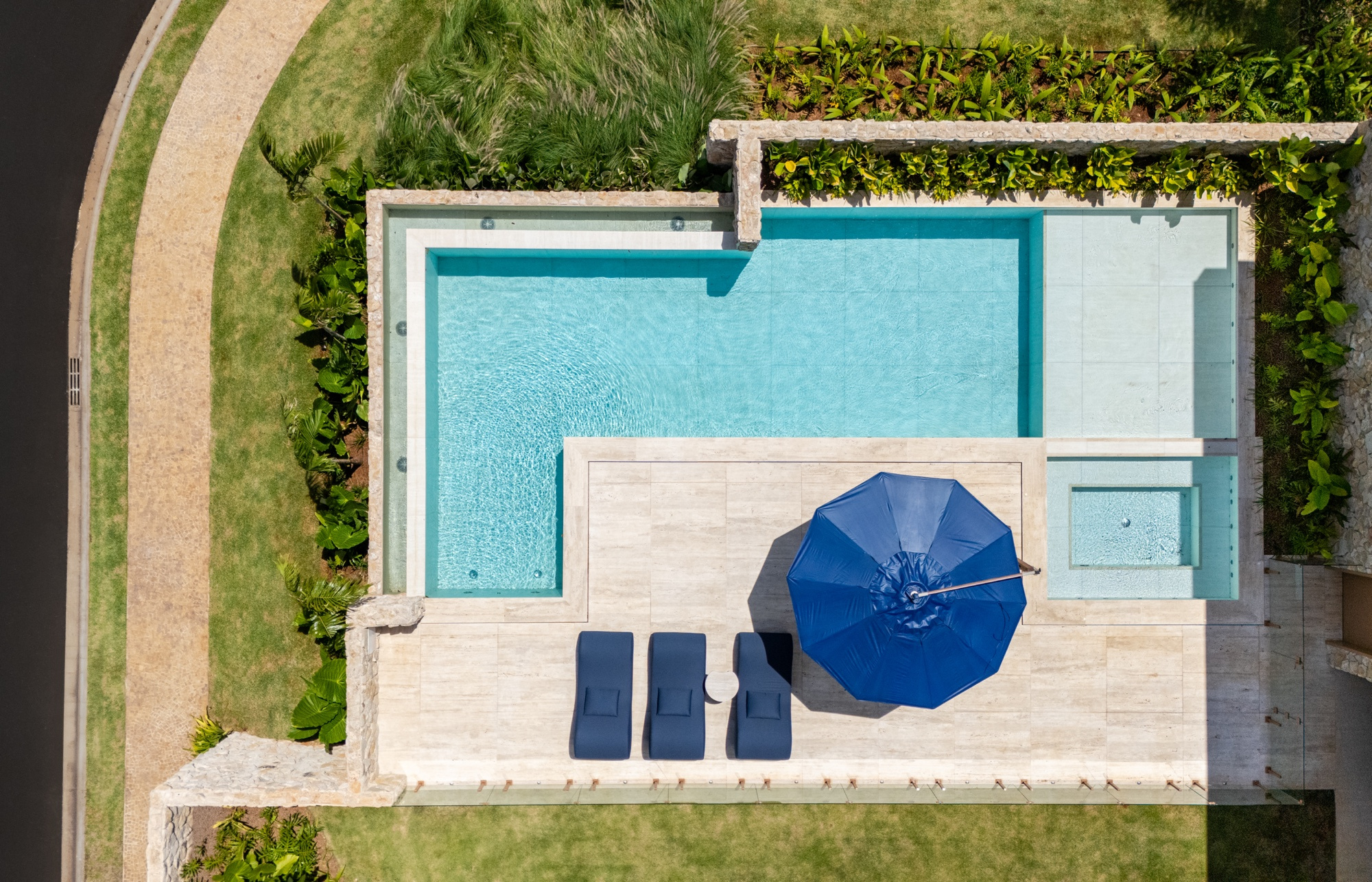 Vista aérea da piscina com guarda-sol azul, deck de pedra, jardineiras de folhagem nas bordas e gramado ao redor