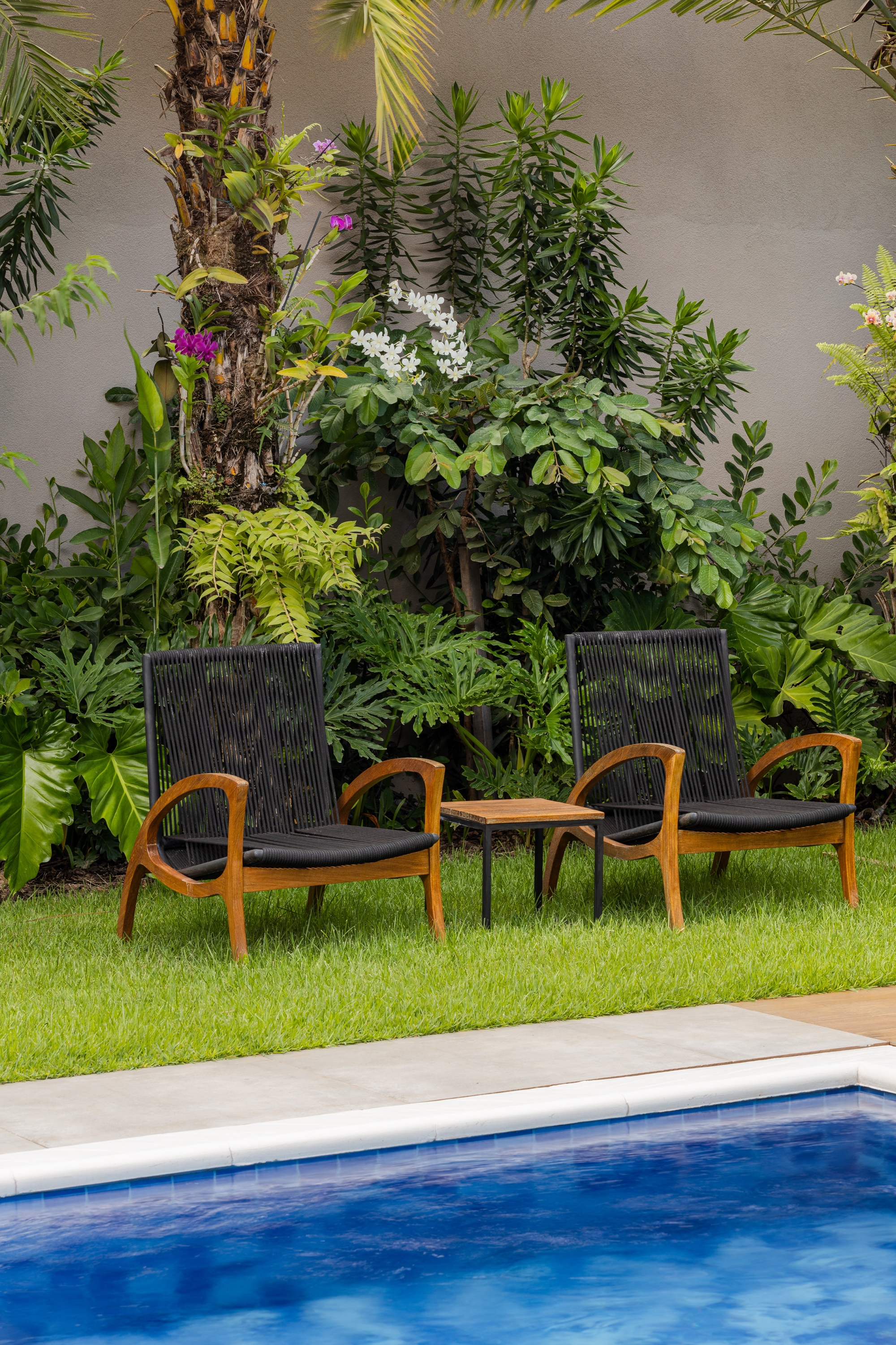 Duas poltronas de madeira e corda com mesinha lateral sobre o gramado à beira da piscina, diante de canteiro tropical com orquídeas roxas e brancas.