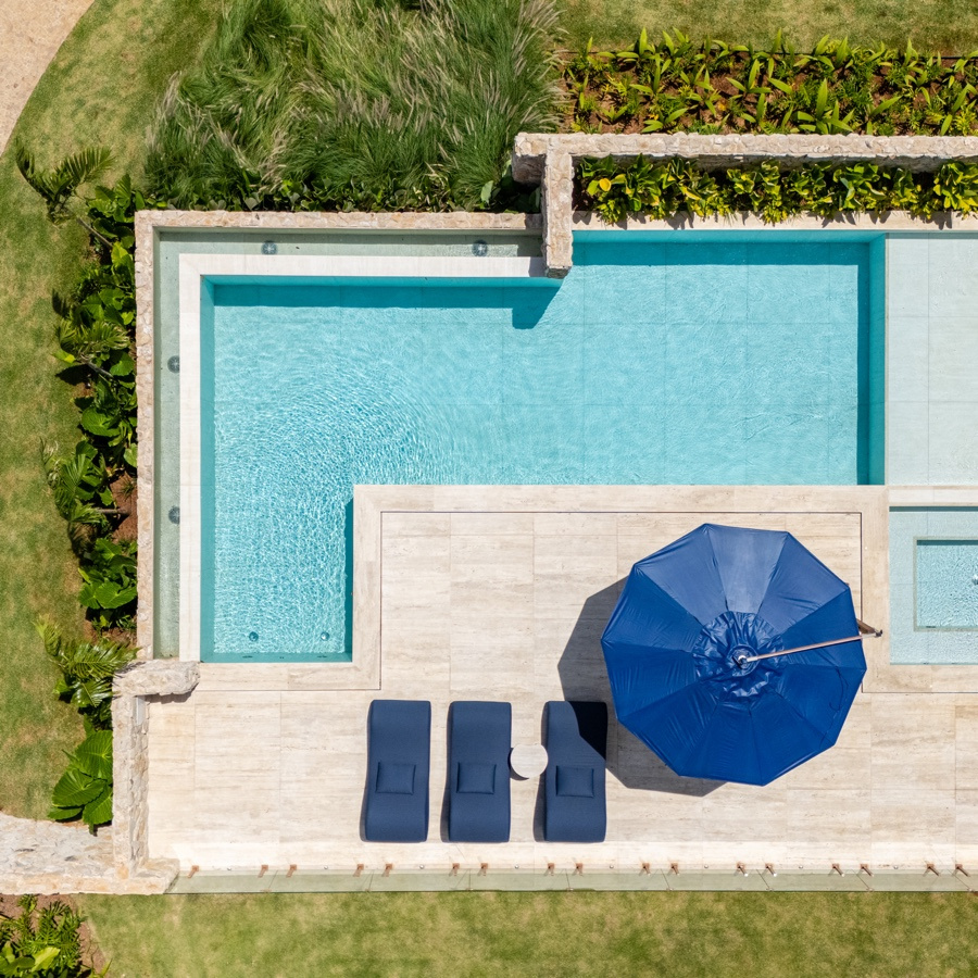Vista aérea da piscina com guarda-sol azul, deck de pedra, jardineiras de folhagem nas bordas e gramado ao redor