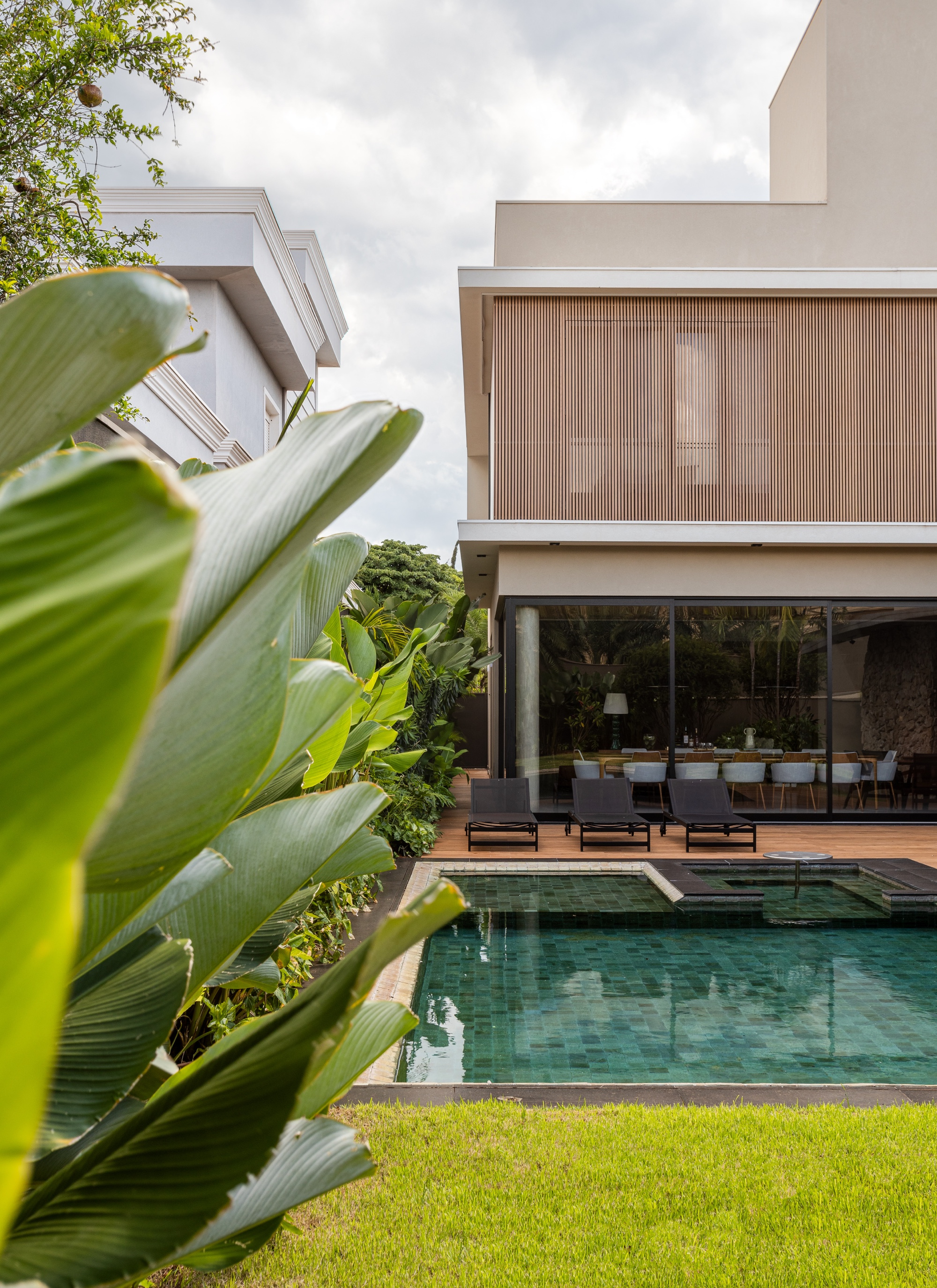 Folhagem de folhas grandes em primeiro plano, piscina ao centro e fachada dos fundos da casa com painel de brise de madeira.