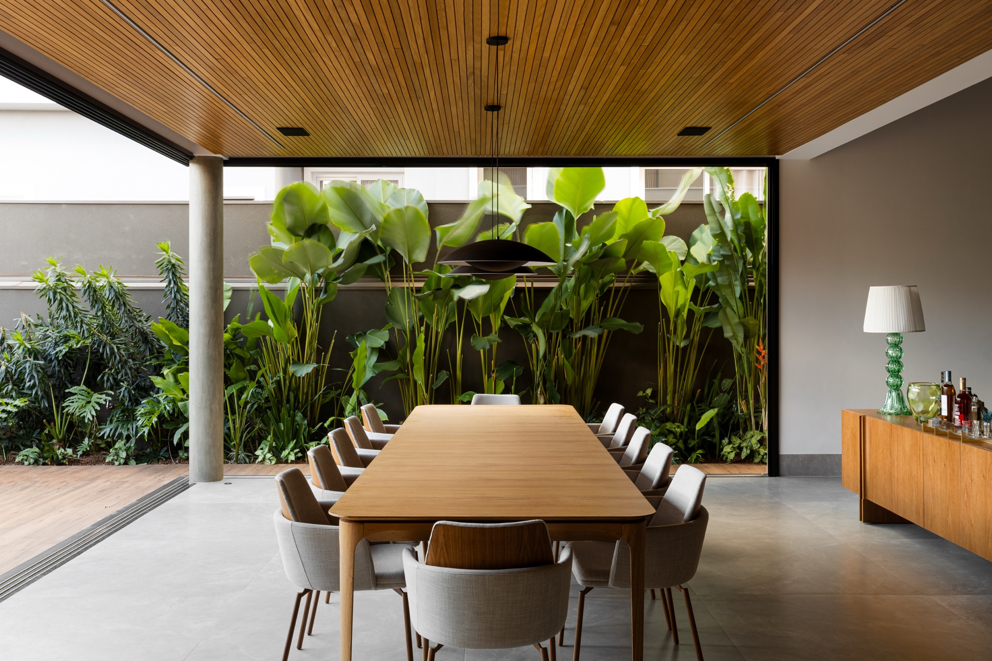 Mesa de jantar longa diante de jardim interno de folhagens altas e verdes, emolduradas pela abertura envidraçada.